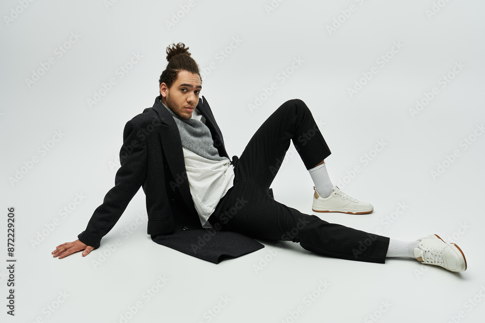 A stylish African American male model wears an elegant coat and sits on a grey background.