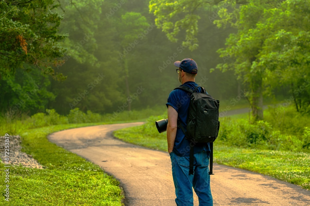 Fototapeta premium Man with a backpack and camera lens walking on a forest path surrounded by lush greenery