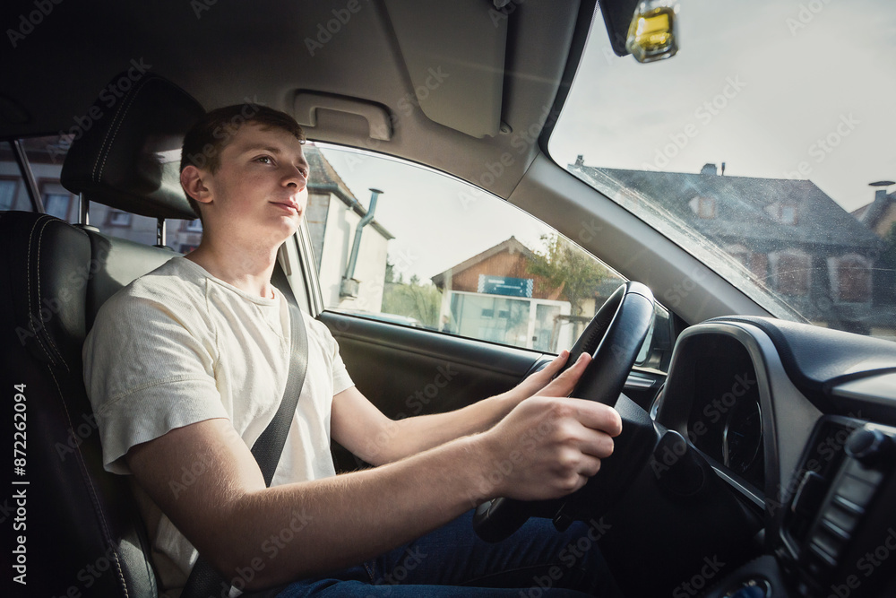 Fototapeta premium Side view young man driving car confident. Teenager driver enjoying the ride on the city street, keeps hands on the steering wheel looking focused at traffic