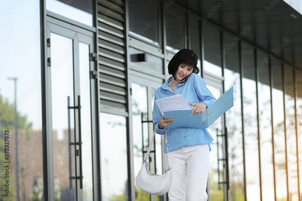Multitasking busy young business woman communicates by smartphone during walks reading and dropping important finance documents by the office buildings outdoors. Stress resistance and business concept Multitasking busy young business woman communicates by smartphone during walks reading and dropping important finance documents by the office buildings outdoors. Stress resistance and business concept