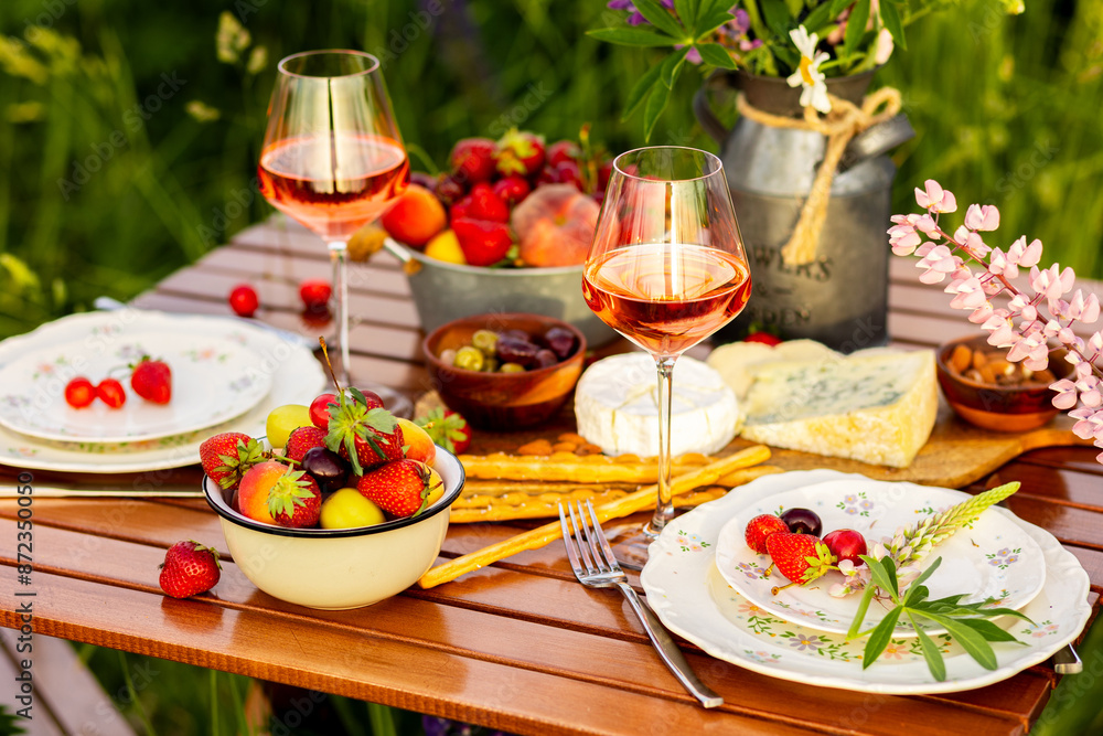 Romantic table decor for a loving couple on blooming meadow with purple lupines. Two glasses of wine, flowers in a vase, silverware, fruits, wooden furniture. Sunset, golden hour, atmospheric light Romantic table decor for a loving couple on blooming meadow with purple lupines. Two glasses of wine, flowers in a vase, silverware, fruits, wooden furniture. Sunset, golden hour, atmospheric light