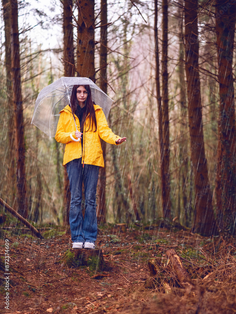 Teenager girl in yellow jacket, blue jeans and translucent umbrella standing on a tree trunk checking out rain. Trip to a forest park. Explore nature theme. Selective focus.