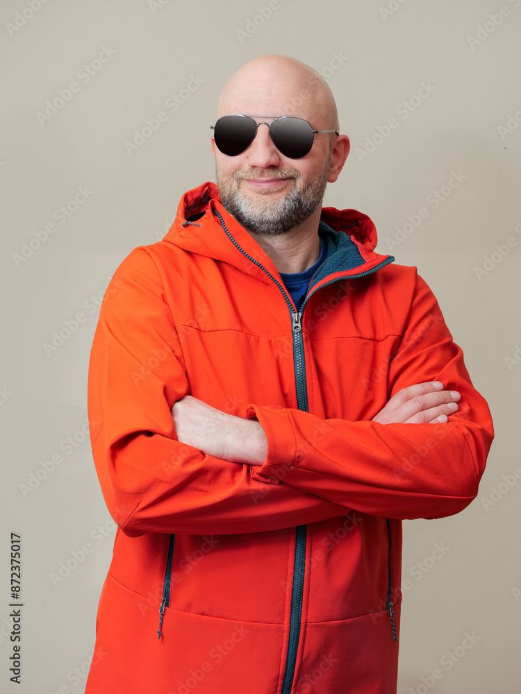 A man in a red jacket hands crossed. The jacket is made of a waterproof material and has a hood and made for hard outdoor conditions. The model is bald, happy and content in dark glasses. Studio shot A man in a red jacket hands crossed. The jacket is made of a waterproof material and has a hood and made for hard outdoor conditions. The model is bald, happy and content in dark glasses. Studio shot