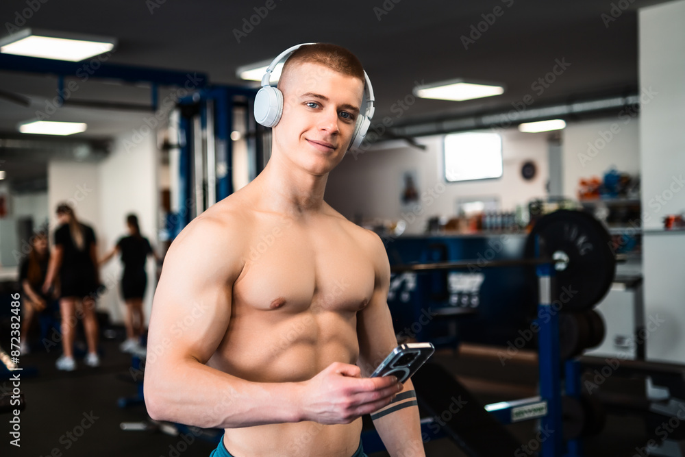 Portrait of a bodybuilder in a gym wears headphones and looks at his phone while resting Portrait of a bodybuilder in a gym wears headphones and looks at his phone while resting