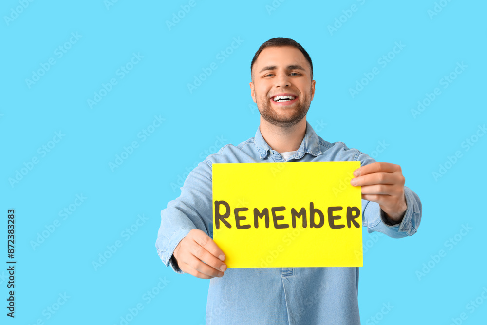 Young man holding paper with word REMEMBER on blue background Young man holding paper with word REMEMBER on blue background