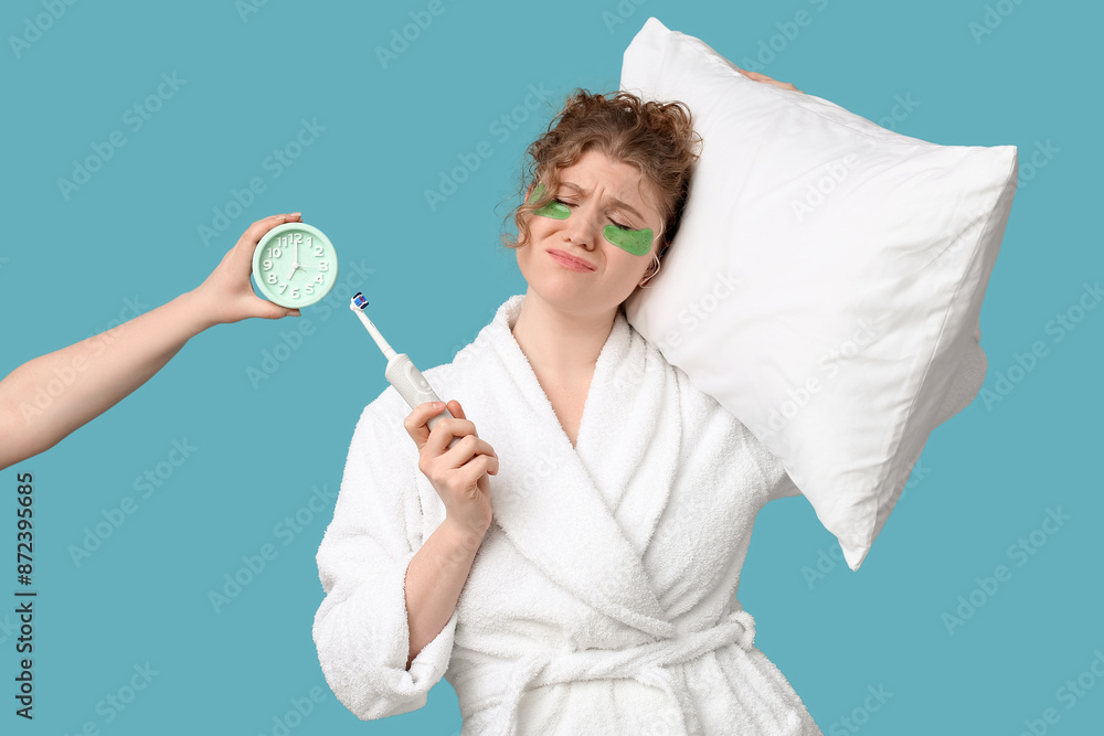 Young sleepy woman with electric toothbrush, pillow and hand holding alarm clock on blue background Young sleepy woman with electric toothbrush, pillow and hand holding alarm clock on blue background