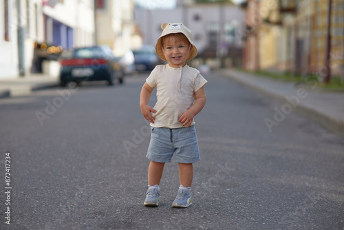 A small handsome boy dressed in a bold T-shirt, shorts and a panama hat is walking around the city. Smiles and poses for the camera.