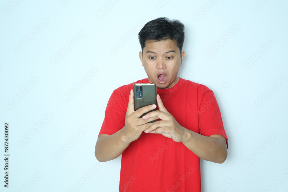 Asian man showing shocking expression looking at smartphone. Wearing red t-shirt Asian man showing shocking expression looking at smartphone. Wearing red t-shirt
