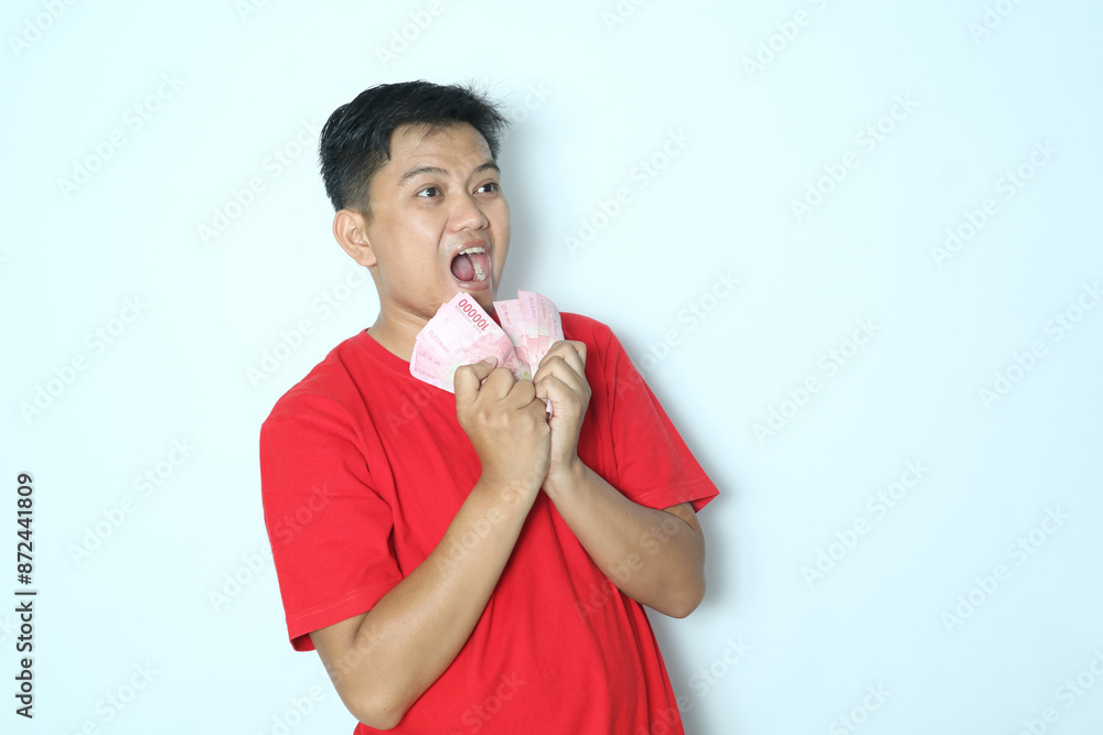 Asian man showing happy face expression while holding paper money with both hands and looking to the side. Wearing red t-shirt Asian man showing happy face expression while holding paper money with both hands and looking to the side. Wearing red t-shirt