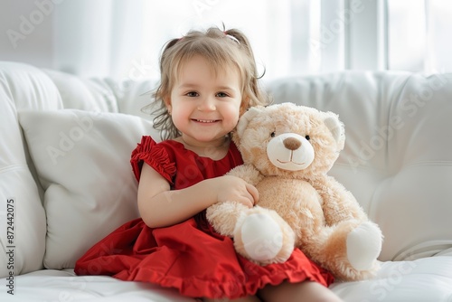 Cute little girl in a red dress smiling and holding a teddy bear, sitting on a white couch, soft natural light from the window.