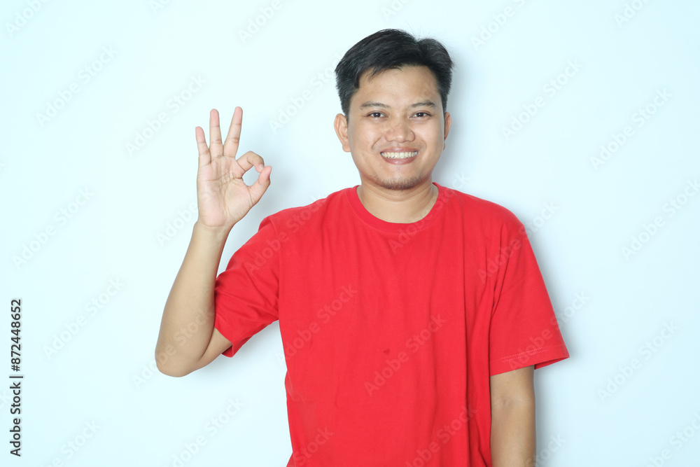 Young Asian man smiling friendly expression while giving the right finger sign OK. Wearing red t-shirt Young Asian man smiling friendly expression while giving the right finger sign OK. Wearing red t-shirt