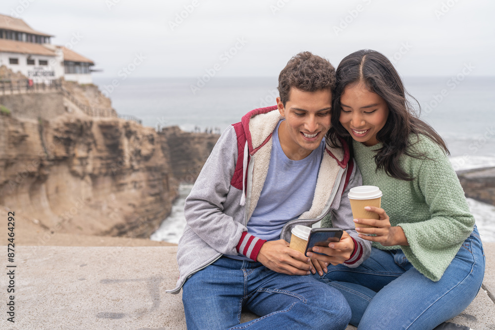 Fototapeta premium Couple drinking coffee and using mobile next to the sea