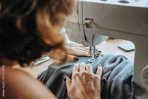 Rear view of a woman seamstress who sits at a sewing machine and sews summer pants from light gray fabric. The work of a professional tailor in an atelier. Tailoring of clothes to order. photo