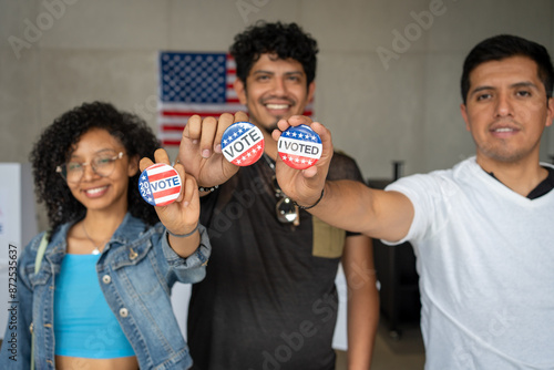 group of Hispanic smiling people holding 2024 Vote election I voted buttons or pins at a US election polling station. In the background, american flag at the background, concept of latin, latino vote