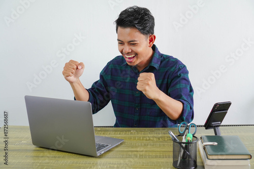 Happy young asian man sit work at wooden desk with pc laptop. Achievement business career lifestyle concept. doing winner gesture while looking at the laptop screen