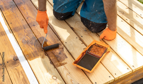 A person applies stain to a wooden deck using a roller. The wood is already partially stained, showcasing the transformation process. photo