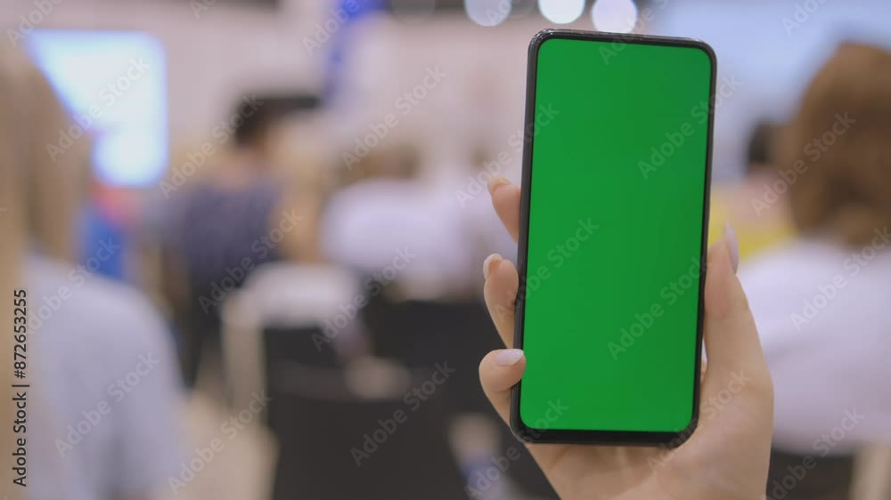 A person holding a smartphone with a green screen in a crowded room, ideal for mockup and presentation use. A person holding a smartphone with a green screen in a crowded room, ideal for mockup and presentation use.