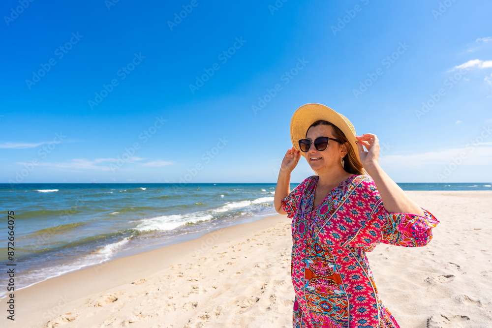 Beach holidays on Baltic Sea. Smiling young plus size woman wearing fashionable multicolored pink dress with hat standing on sunny sandy beach on windy day. Front view Beach holidays on Baltic Sea. Smiling young plus size woman wearing fashionable multicolored pink dress with hat standing on sunny sandy beach on windy day. Front view