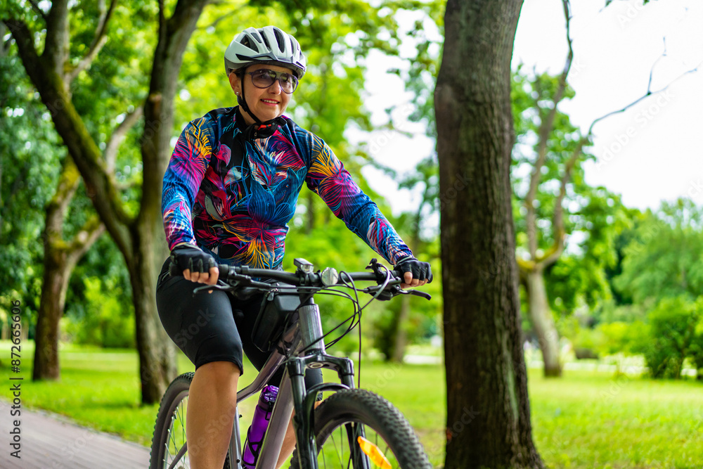 Cycling training in city. Young joyful woman in white helmet and colorful blouse and black shorts riding bike on bike path in city. Playing sports outdoors in spring. Healthy lifestyle Cycling training in city. Young joyful woman in white helmet and colorful blouse and black shorts riding bike on bike path in city. Playing sports outdoors in spring. Healthy lifestyle