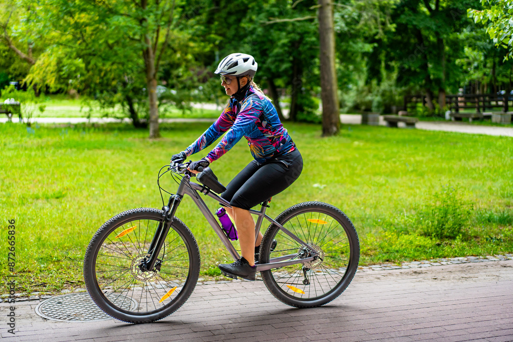 Cycling training in city. Young joyful woman in white helmet and colorful blouse and black shorts riding bike on bike path in city. Playing sports outdoors in spring. Healthy lifestyle Cycling training in city. Young joyful woman in white helmet and colorful blouse and black shorts riding bike on bike path in city. Playing sports outdoors in spring. Healthy lifestyle
