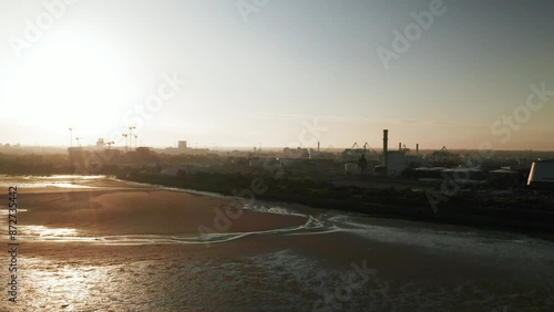 Aerial footage at sunrise capturing the serene Sandymount Beach in Dublin, with the industrial skyline of Dublin Port, including cranes and smoke stacks, illuminated by the early morning light.