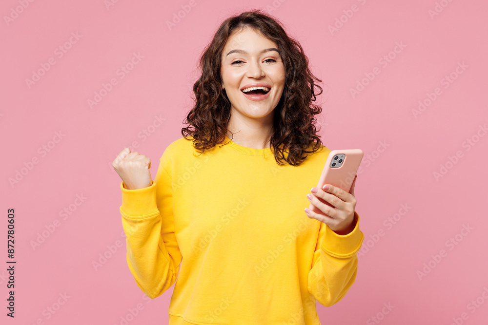Young happy fun cool woman she wears yellow sweatshirt casual clothes hold in hand use mobile cell phone do winner gesture isolated on pastel light pink background studio portrait. Lifestyle concept. Young happy fun cool woman she wears yellow sweatshirt casual clothes hold in hand use mobile cell phone do winner gesture isolated on pastel light pink background studio portrait. Lifestyle concept.