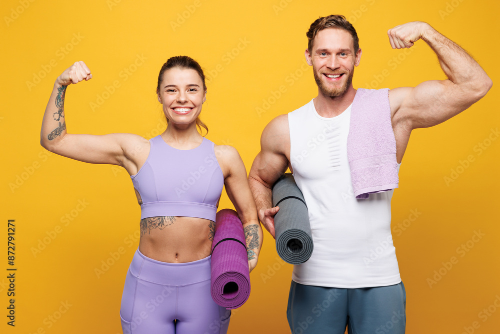 Young strong fitness trainer instructor sporty two man woman wear blue clothes spend time in home gym hold yoga mat caremat show muscles isolated on plain yellow background. Workout sport fit concept. Young strong fitness trainer instructor sporty two man woman wear blue clothes spend time in home gym hold yoga mat caremat show muscles isolated on plain yellow background. Workout sport fit concept.