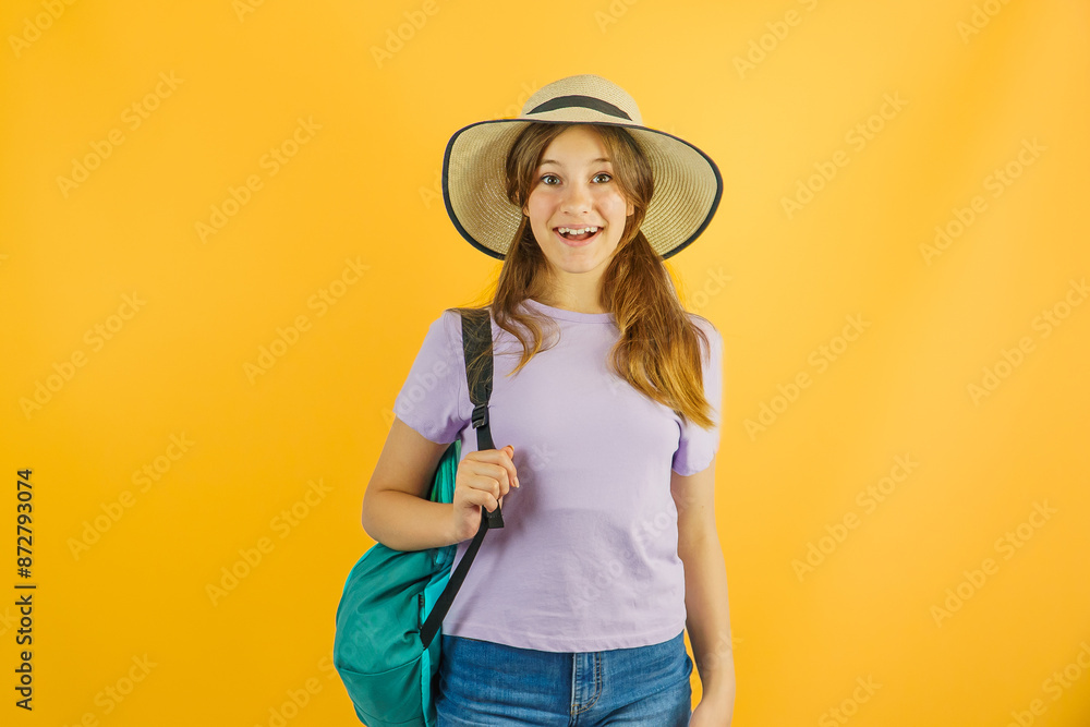 Happy little cute girl traveler carries a hiking backpack, travels on a hike, stands on a yellow background, wears a stylish hat, has a joyful expression on her face, loves to travel