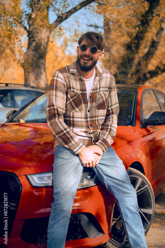 Smiling young Caucasian man leaning on a car, travel enthusiast sharing stories about trips and nature adventures. photo