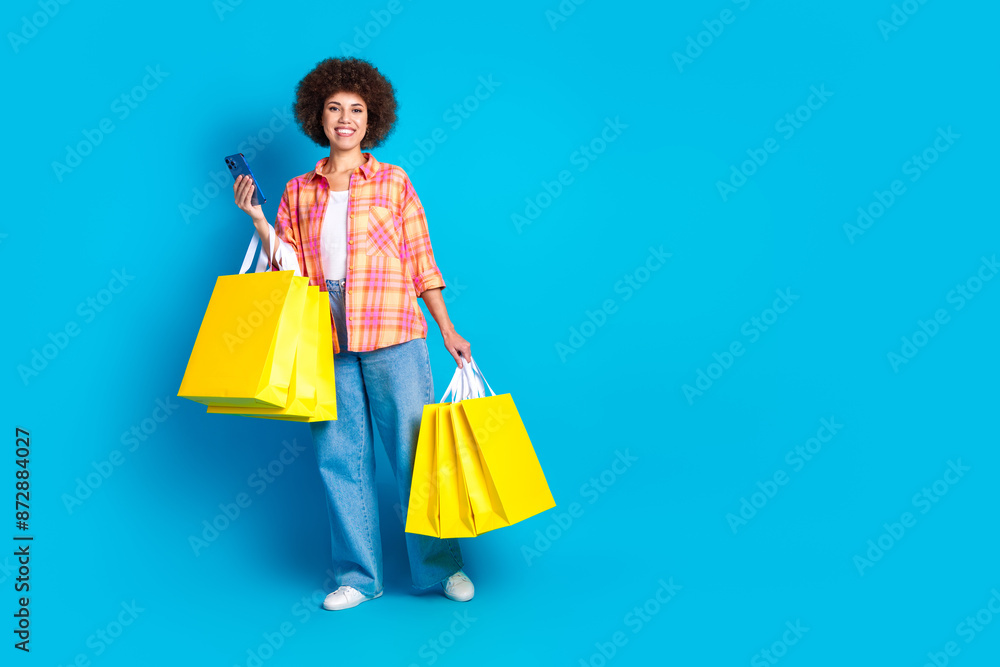 Full size photo of attractive young woman hold device shopping bags dressed stylish checkered clothes isolated on blue color background Full size photo of attractive young woman hold device shopping bags dressed stylish checkered clothes isolated on blue color background