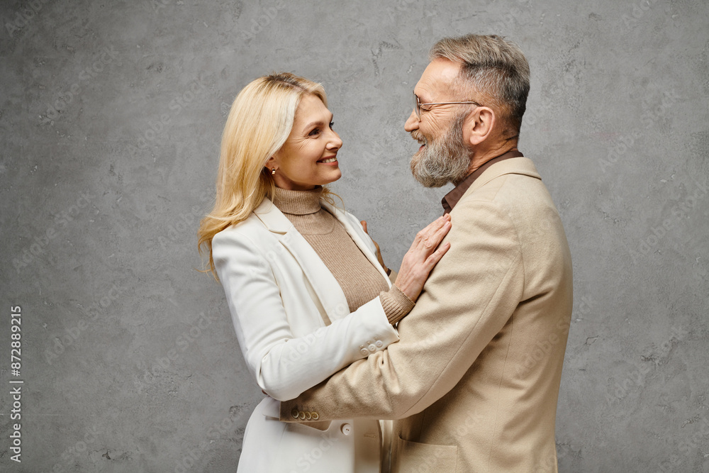 Elegant, mature man and woman in stylish attire pose together against a gray backdrop. Elegant, mature man and woman in stylish attire pose together against a gray backdrop.