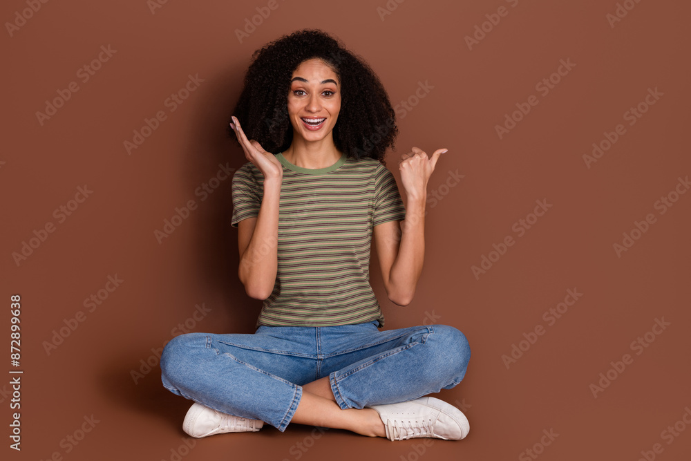 Full length photo of cheerful pretty lady dressed khaki t-shirt sitting legs crossed shwoing thumb empty space isolated brown color background Full length photo of cheerful pretty lady dressed khaki t-shirt sitting legs crossed shwoing thumb empty space isolated brown color background