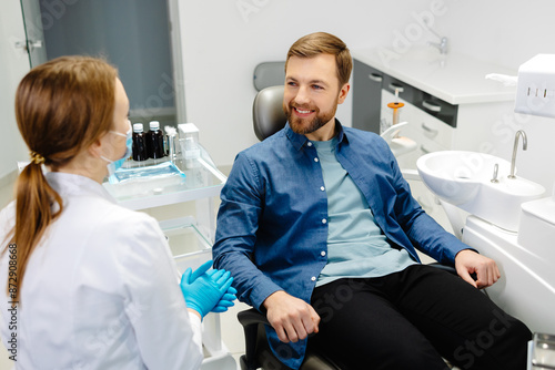 Blonde female dentist in dental office talking with male patient and preparing for treatment. Handsome bearded man in dentist chair looking at his doctor with smile
