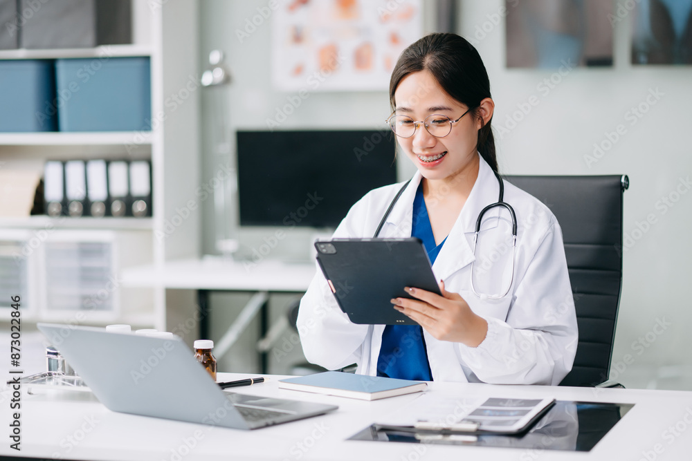 Confident young Asian female doctor in white medical uniform sit at desk working on computer. Smiling use laptop write in medical journal Confident young Asian female doctor in white medical uniform sit at desk working on computer. Smiling use laptop write in medical journal