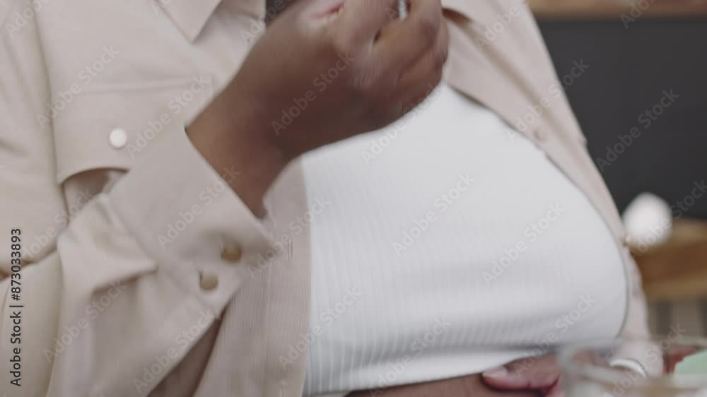 Medium tilt of young pregnant African American woman in casual outfit sitting at table in kitchen taking supplement pill, drinking water and gently caressing abdomen Medium tilt of young pregnant African American woman in casual outfit sitting at table in kitchen taking supplement pill, drinking water and gently caressing abdomen