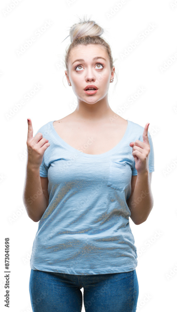 Young blonde woman over isolated background amazed and surprised looking up and pointing with fingers and raised arms. Young blonde woman over isolated background amazed and surprised looking up and pointing with fingers and raised arms.