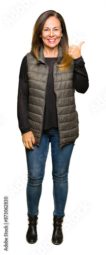 Beautiful middle age woman wearing winter vest smiling with happy face looking and pointing to the side with thumb up.