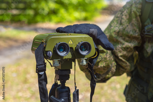 Army rangefinder-angle meter on a tripod. Soldier's hand in a tactical glove. Close-up. Determining the distance to various objects and conducting optical reconnaissance. Blurred background.