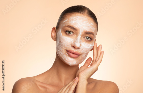 Beautiful young European woman applying facial mask on her face, enjoying skin care and treatment over beige background photo