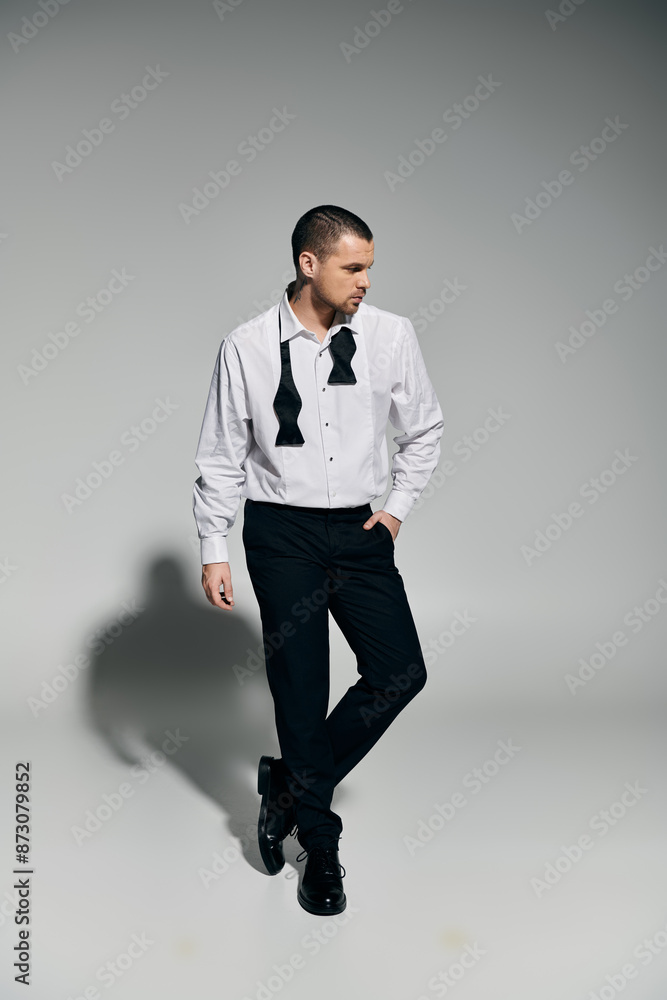 Man in white shirt, black bow tie poses with hand in pocket against grey background.