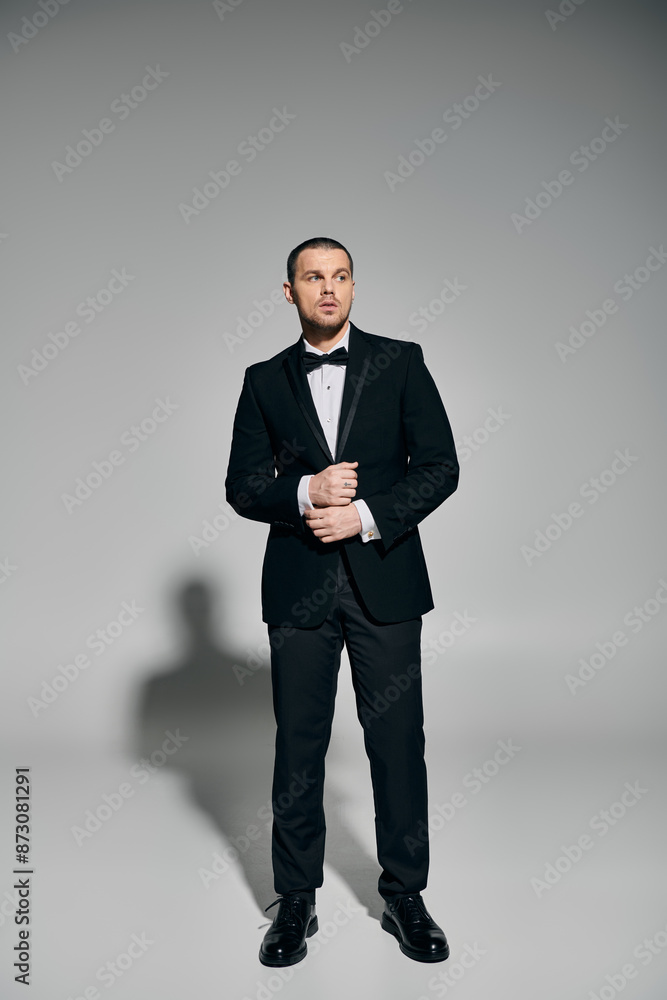 A man in a black tuxedo stands against a grey backdrop, looking up and adjusting his jacket. A man in a black tuxedo stands against a grey backdrop, looking up and adjusting his jacket.