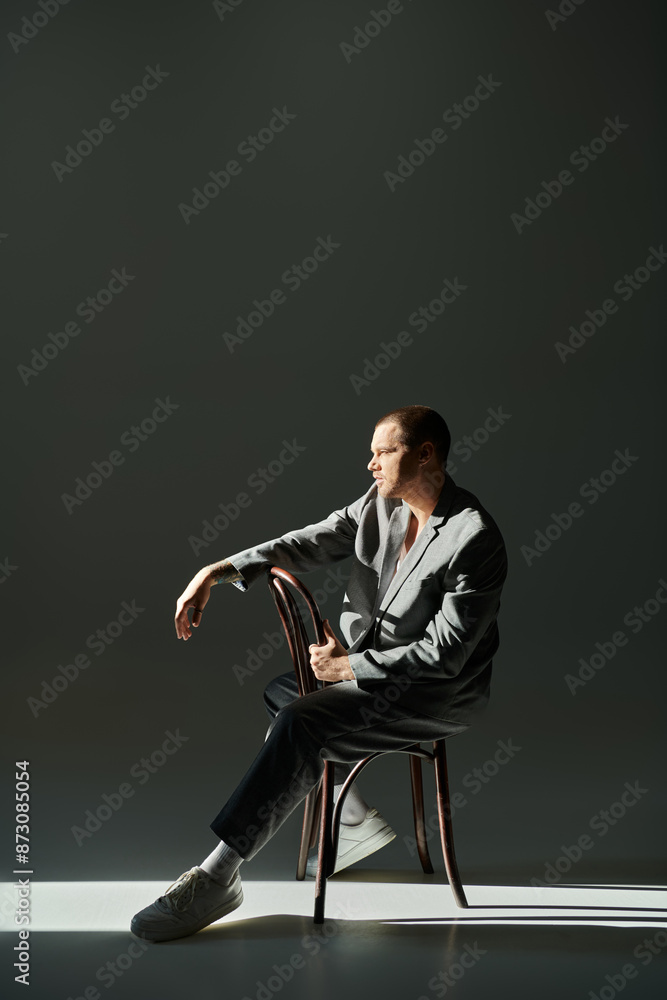A stylish man in a grey suit sits on a wooden chair, gazing off to the side. A stylish man in a grey suit sits on a wooden chair, gazing off to the side.