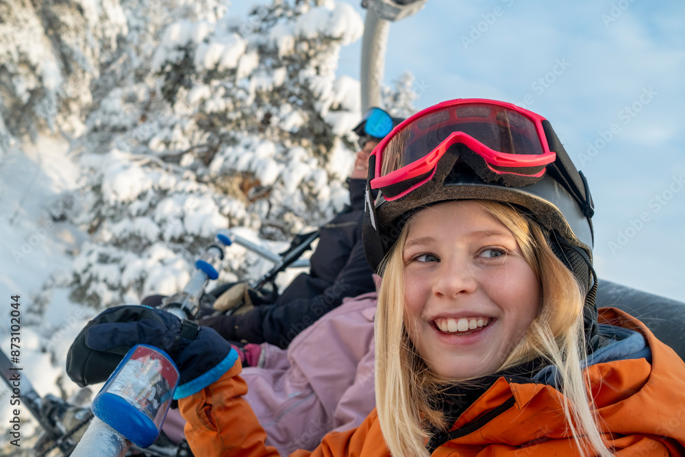 Smiling girl wearing helmet and goggles sitting on ski lift Smiling girl wearing helmet and goggles sitting on ski lift