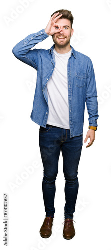 Young handsome blond man wearing casual denim jacket doing ok gesture with hand smiling, eye looking through fingers with happy face.