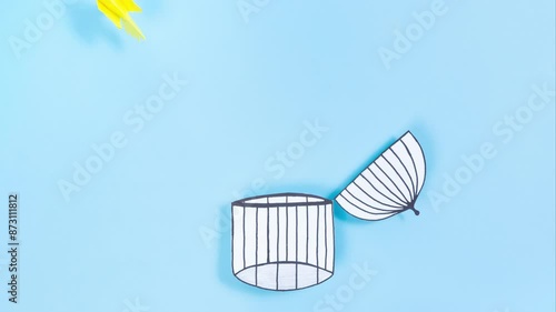 Concept of gaining freedom, removing limiting attitudes, beliefs, liberation from the cage. Yellow bird flies out of the opening cage. Blue background. Flat lay. Stop motion animation.