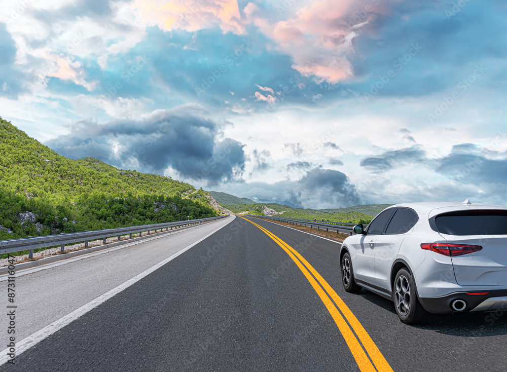A white car drives along the highway against the backdrop of rocky mountains on a sunny day.