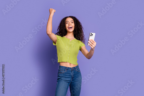 Photo portrait of attractive teen woman hold device winning dressed stylish green clothes isolated on purple color background