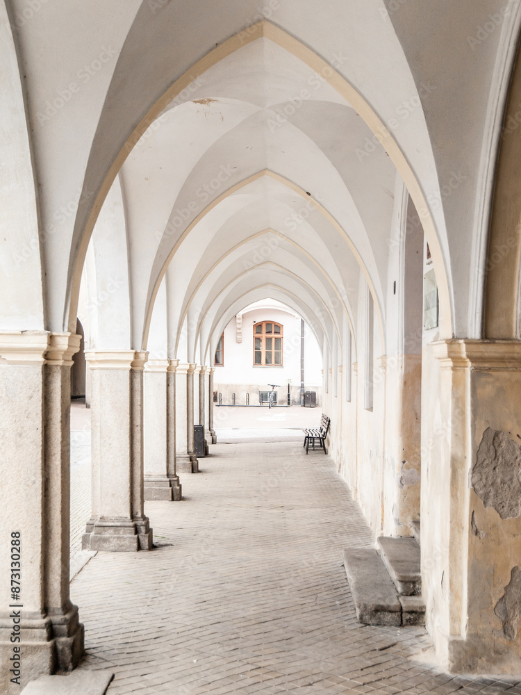 Obraz premium A long, arched walkway with white stone columns leads to a courtyard with a bench in the distance.