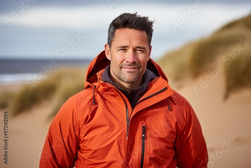 Portrait of a glad man in his 40s wearing a windproof softshell while standing against sandy beach background