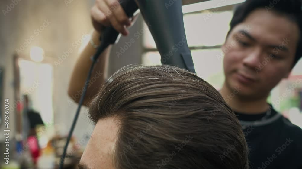 Medium close up of Asian barber doing styling using hairdryer on hair of male client at barbershop Medium close up of Asian barber doing styling using hairdryer on hair of male client at barbershop